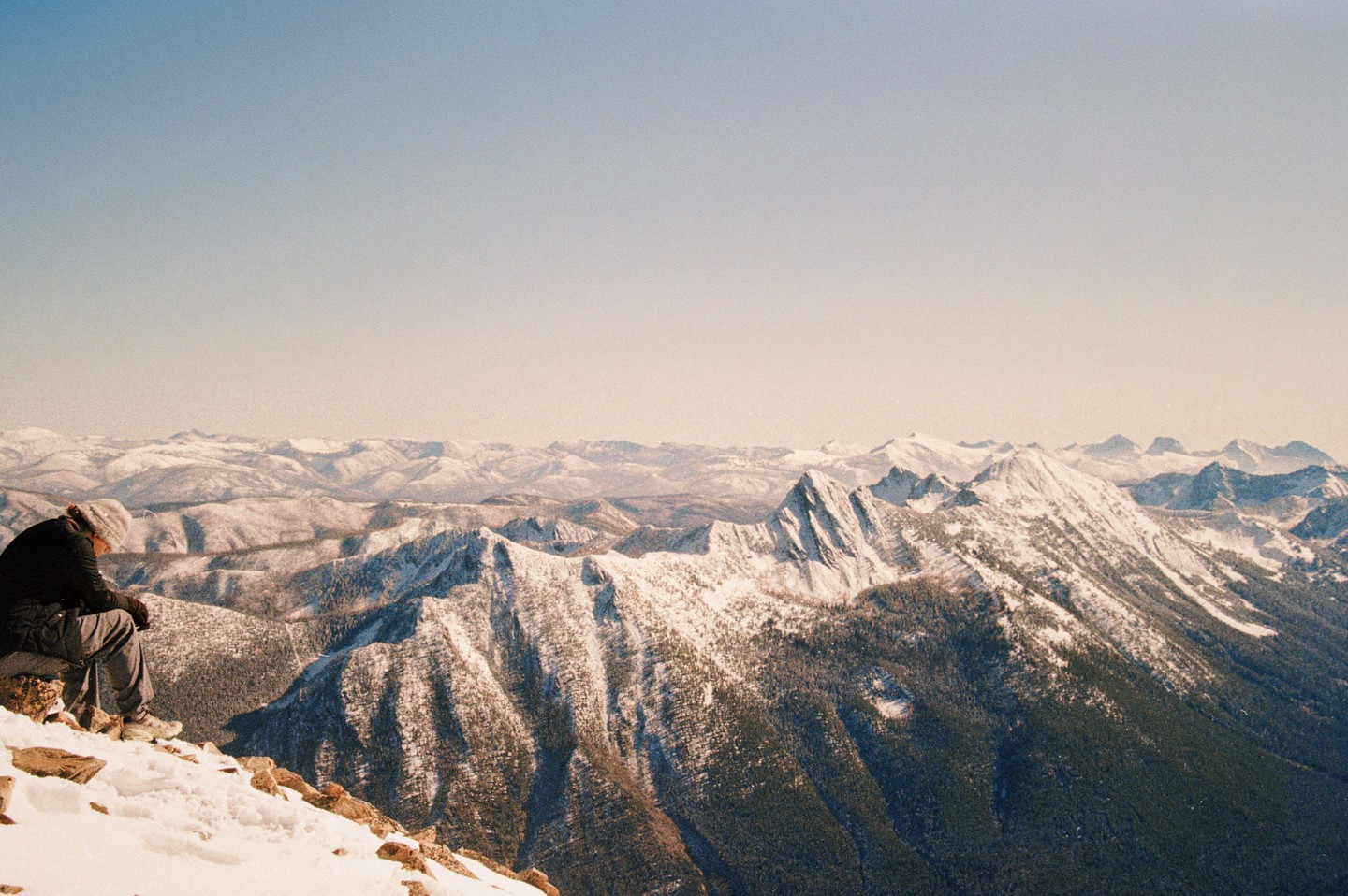 A person sits on a snowy mountain peak overlooking a vast mountain range.