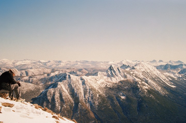 A person sits on a snowy mountain peak overlooking a vast mountain range.