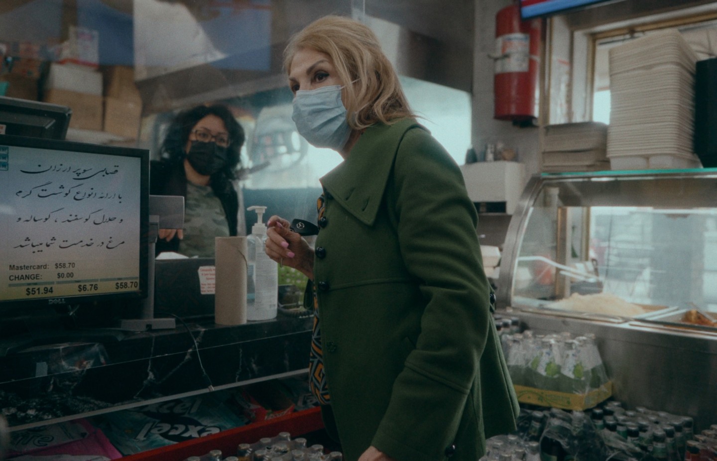 A woman in a green coat and face mask stands at a counter in a store with another woman visible behind a screen.