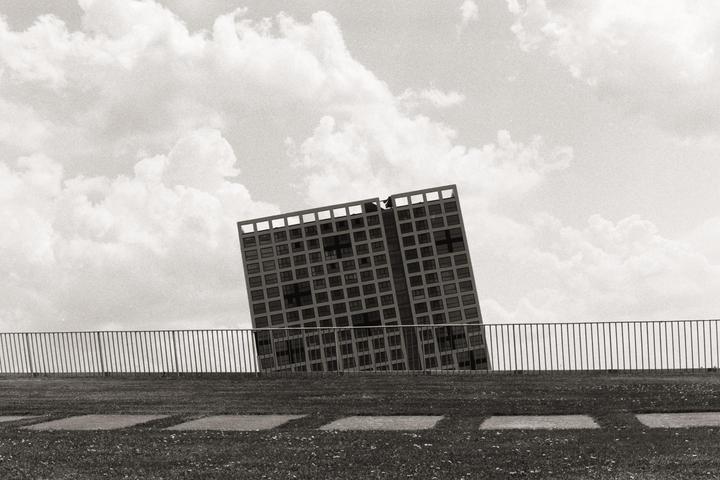 A black and white photo shows a tall building with many windows, appearing to lean to the left, behind a metal fence and a grassy area with square stepping stones.