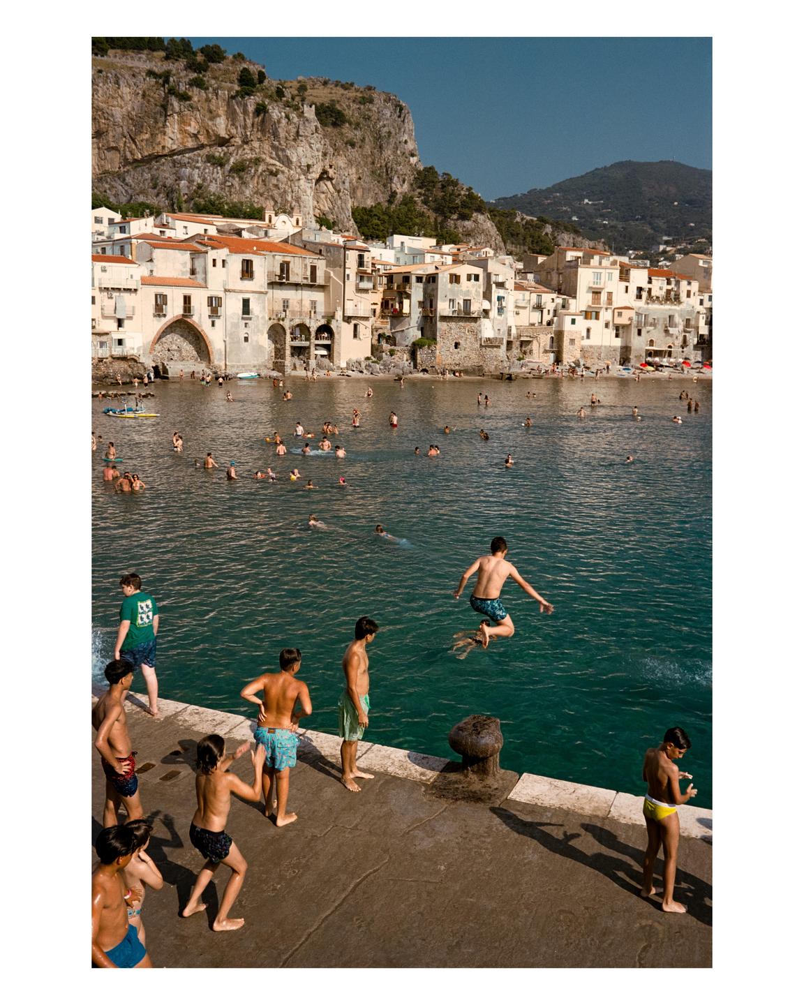 A boy jumps into the water from a concrete pier, while other boys stand on the pier and many people swim in the water in front of a town built into a cliff.