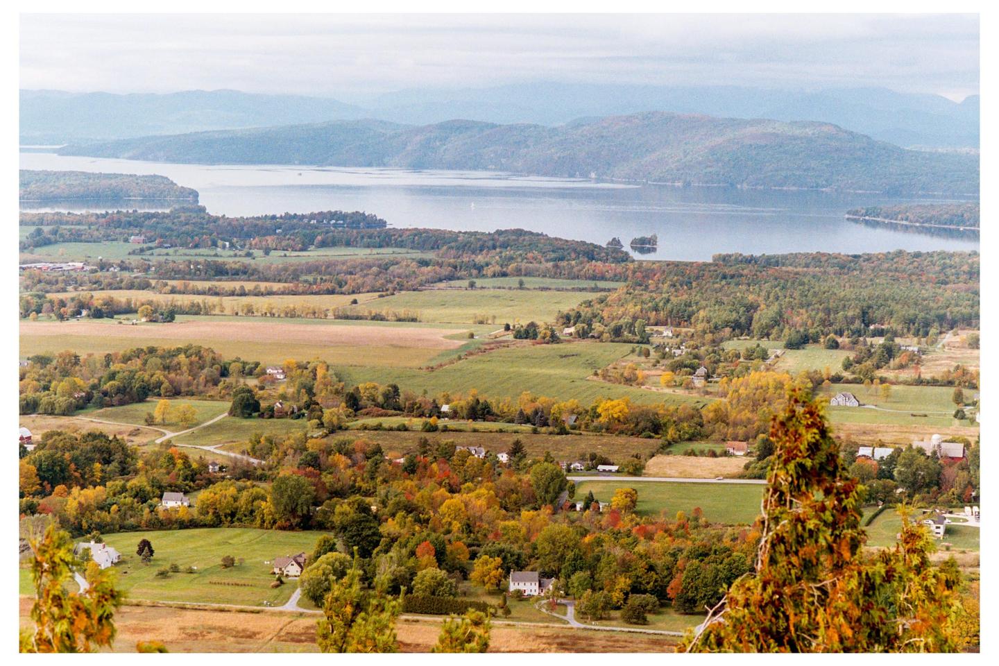 A wide landscape shows a body of water surrounded by hills and fields with autumn foliage.
