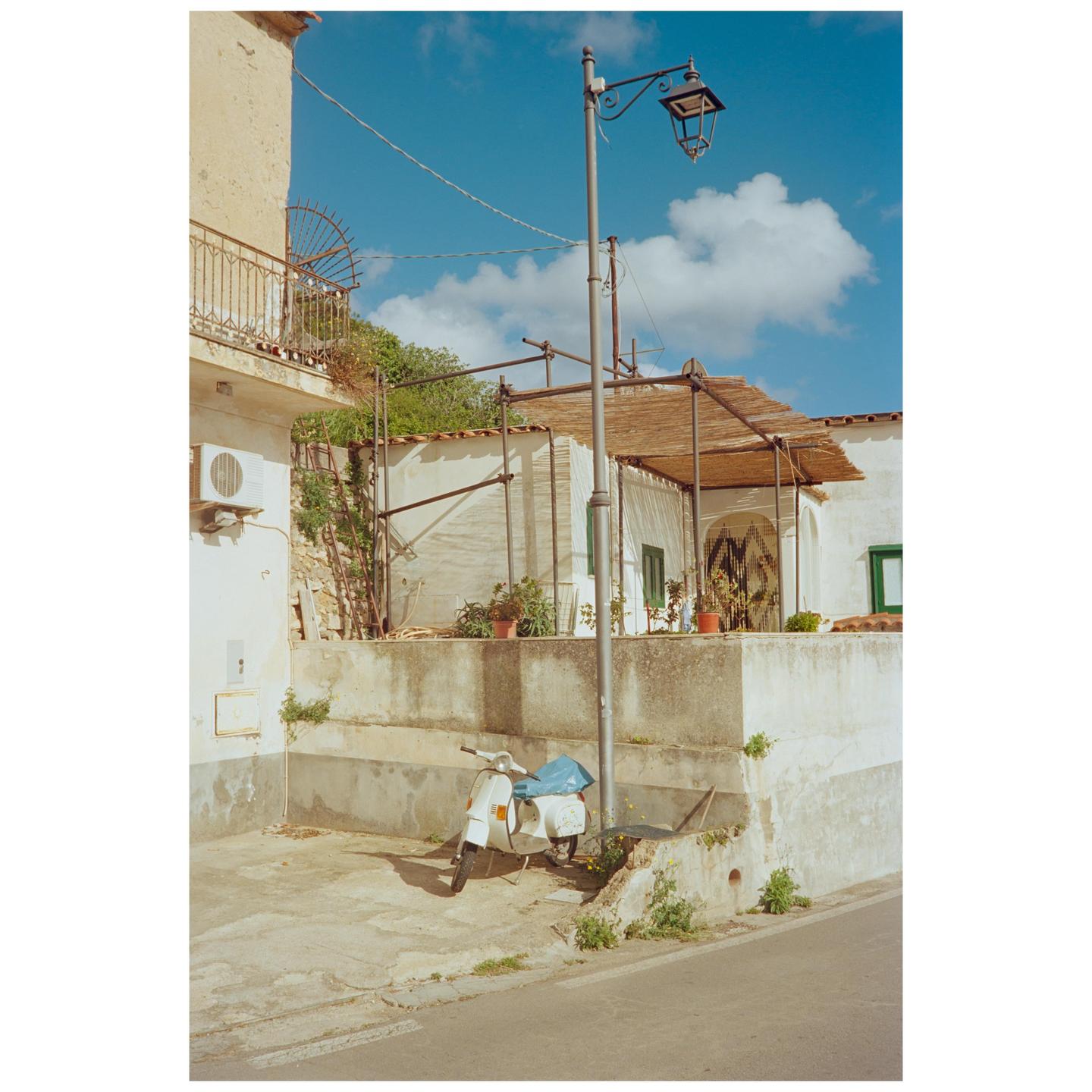 A white scooter is parked on a concrete surface next to a building with a balcony and a street lamp.