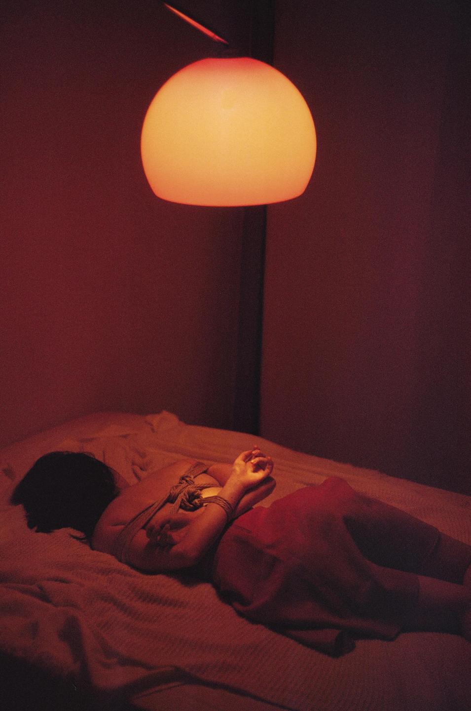 A woman lies on a bed with her hands tied behind her back under a glowing orange lamp.