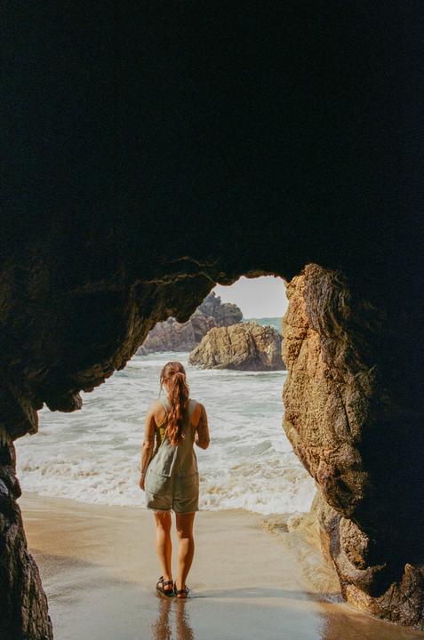 A woman stands on a sandy beach looking out at the ocean through a cave opening.