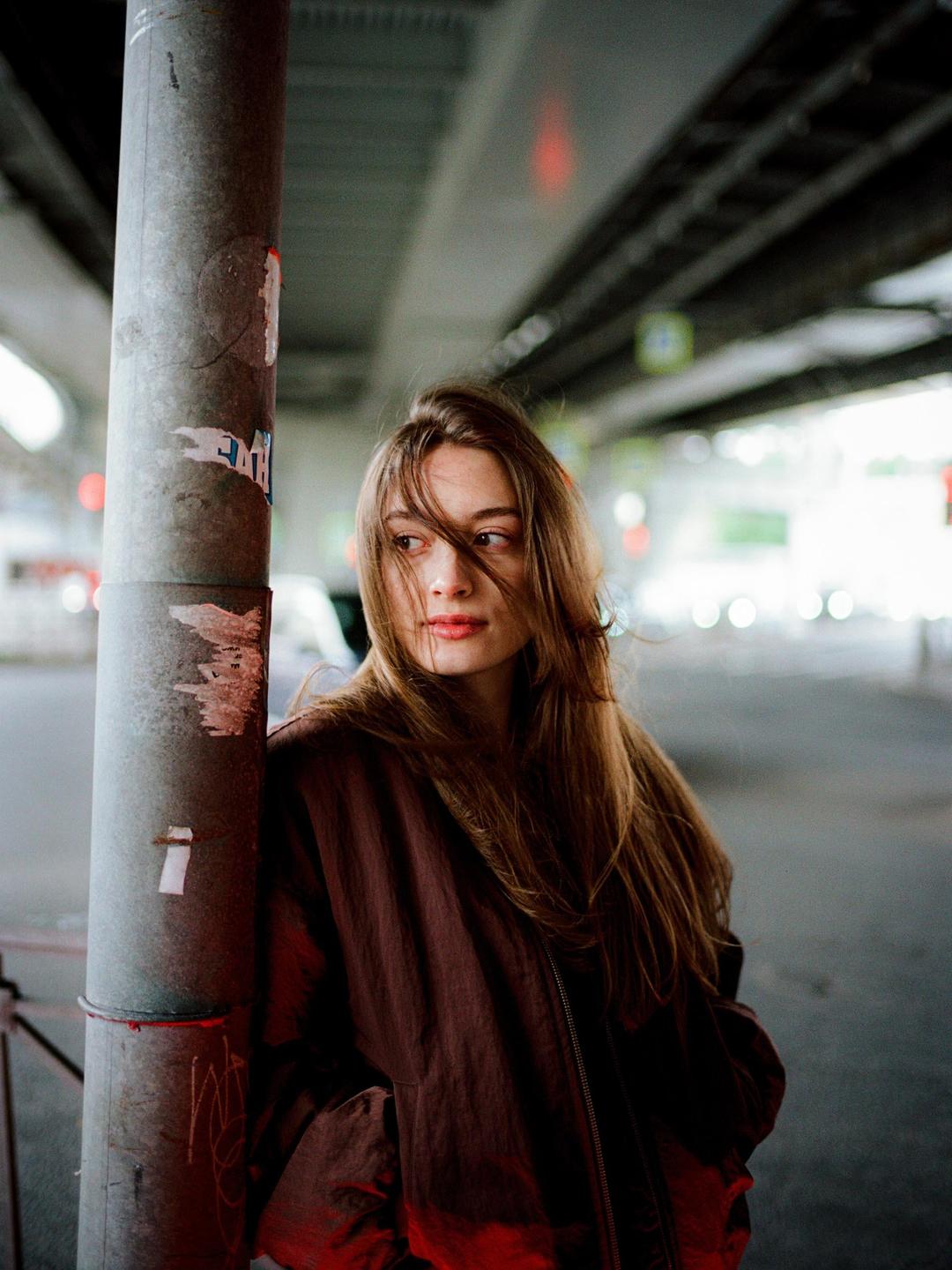 A woman with long hair and a brown jacket leans against a pole under an overpass.