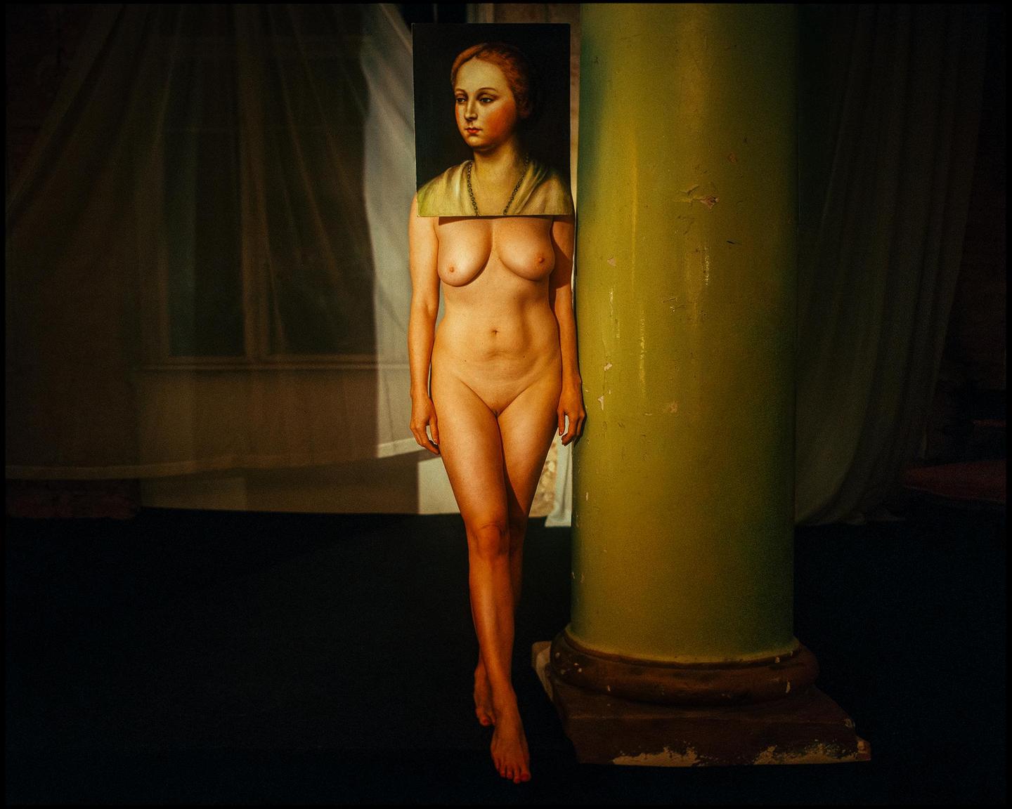 A nude woman with a painting of a woman's head covering her own stands next to a green column.