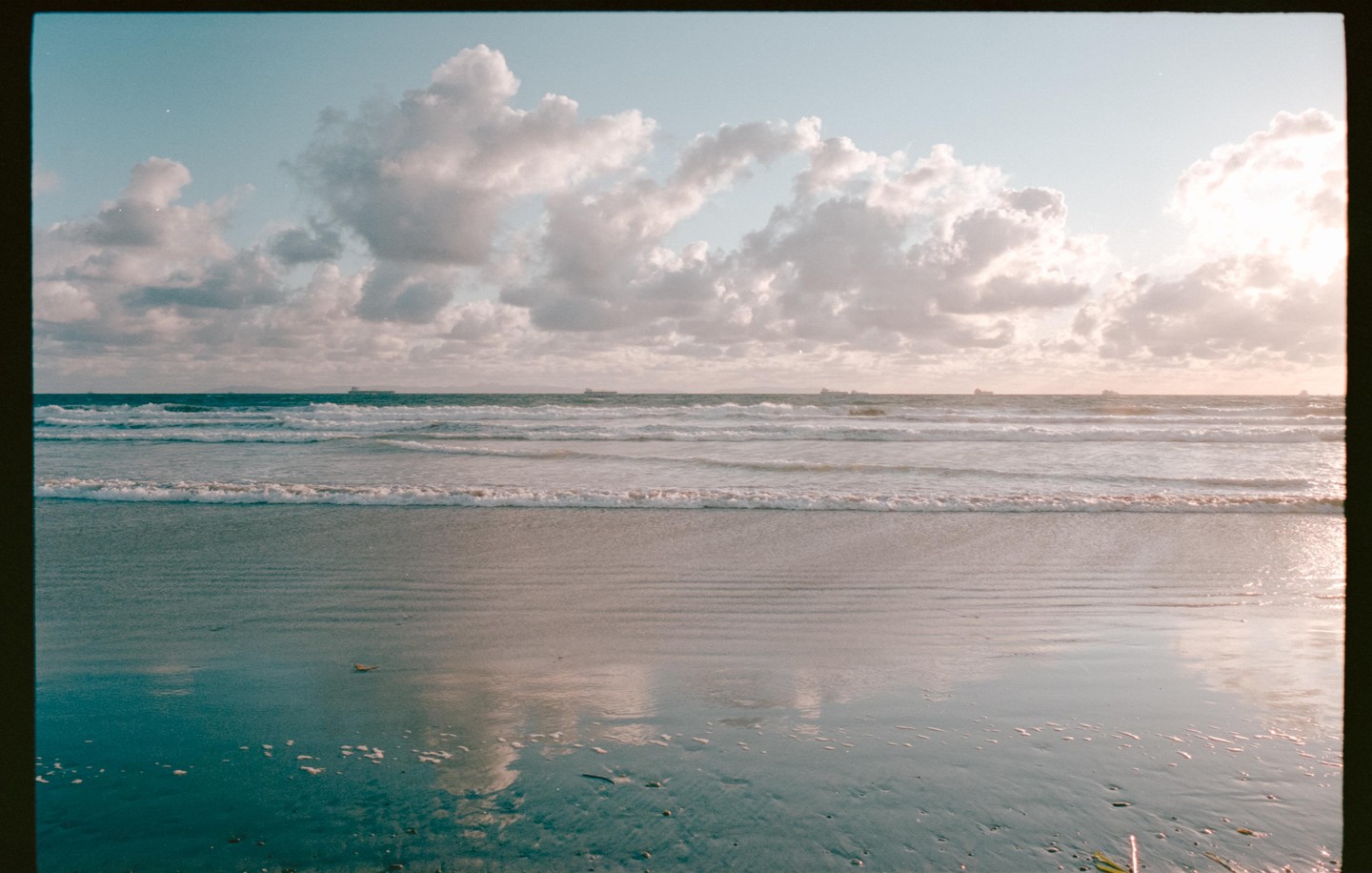 Waves crash on a beach under a cloudy sky with ships visible on the horizon.