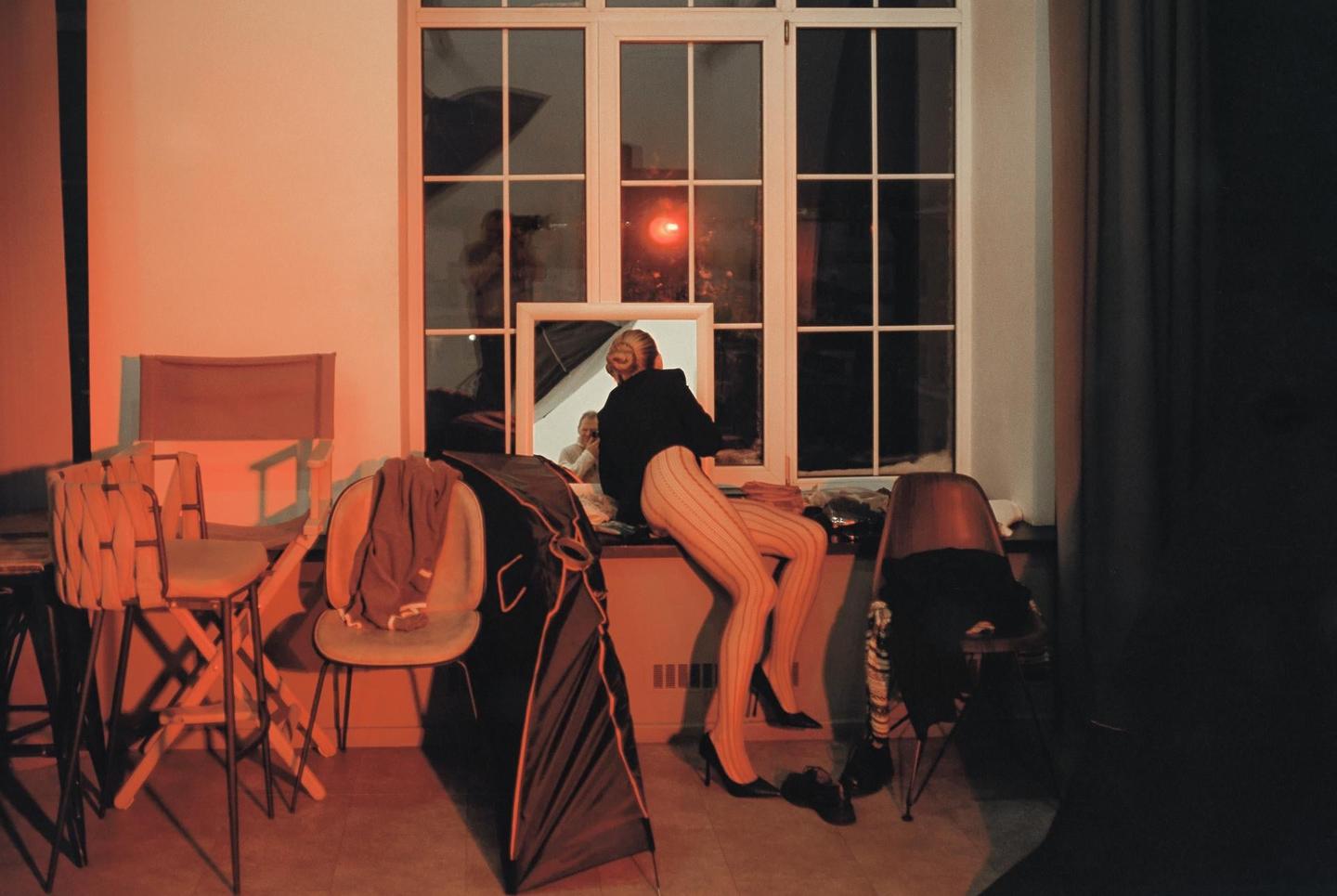 A woman in striped tights and high heels sits on a window ledge, looking into a mirror, in a room with orange lighting.
