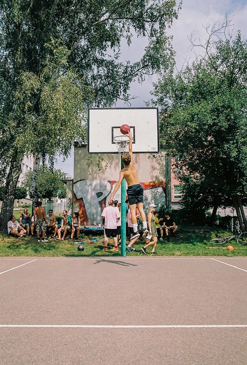 A young man dunks a basketball on an outdoor court while other people watch and relax nearby.
