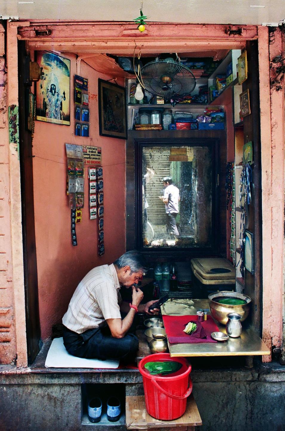 A man sits cross-legged in a small shop, looking at his phone, with a reflection of another man visible in a mirror.