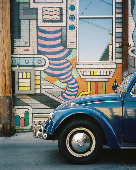 A blue car is parked next to a building with a colorful mural and a window.
