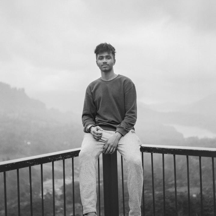 A young man sits on a railing overlooking a mountainous landscape.