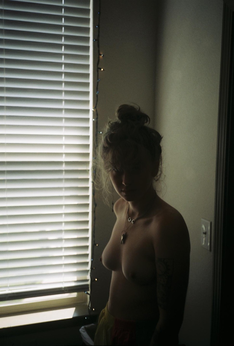 A woman with a top bun and a necklace stands topless next to a window with blinds and string lights.