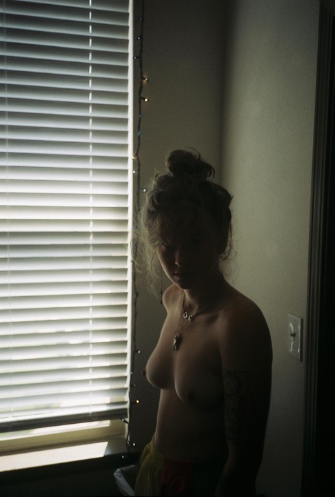 A woman with a top bun and a necklace stands topless next to a window with blinds and string lights.