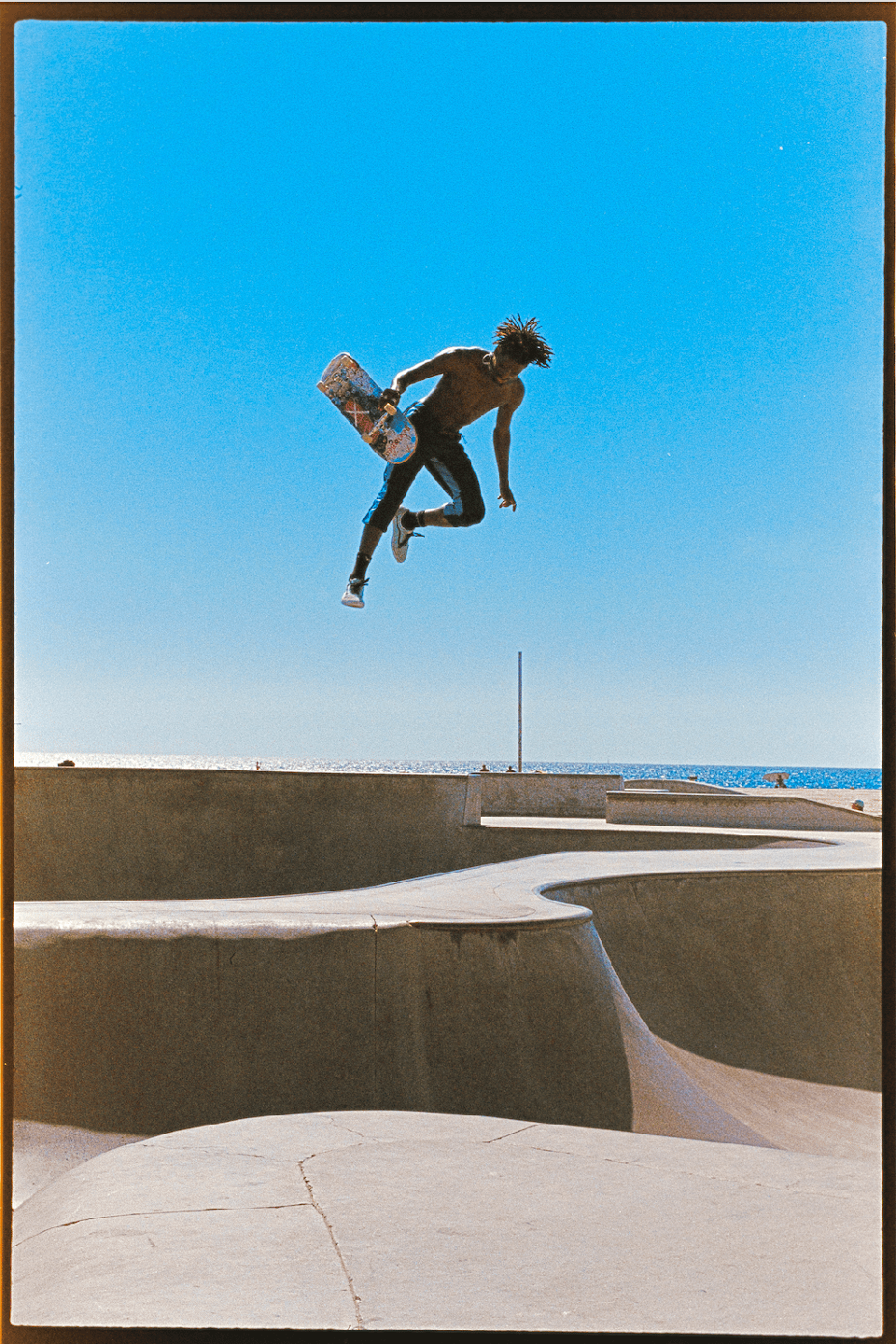A man with a skateboard is captured mid-air above a concrete skatepark with the ocean visible in the background.