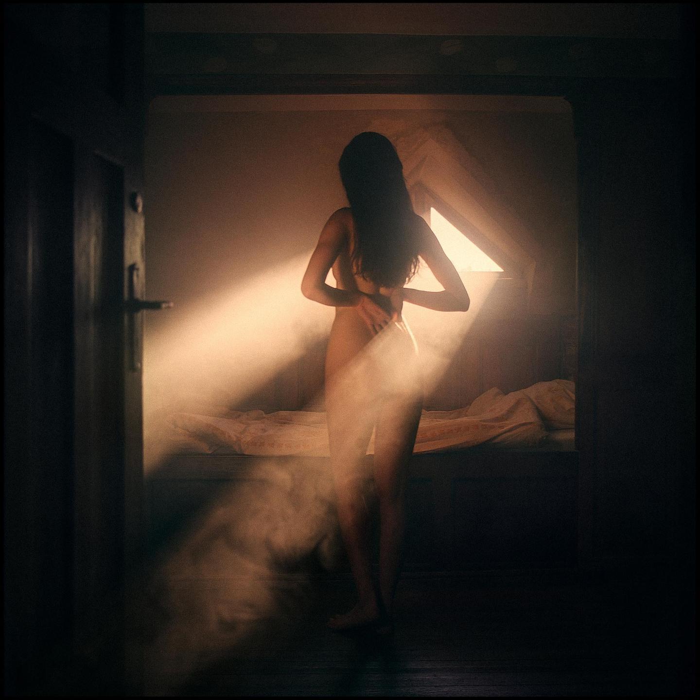 A woman stands in a dimly lit room with a ray of light shining through a window.