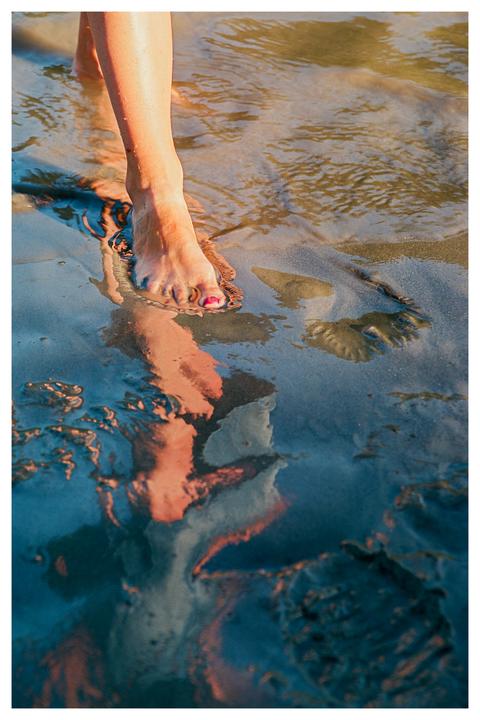 A woman's foot with painted toenails steps into shallow water on a beach during golden hour.