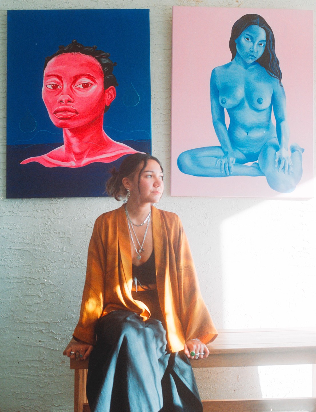 A woman sits on a bench in front of two paintings, one with a red head and the other with a blue nude figure.