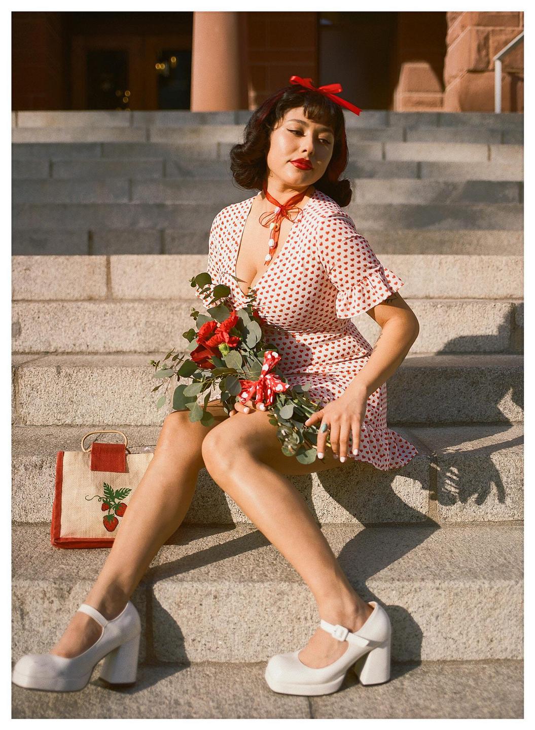 A woman in a heart-patterned dress sits on stone steps, holding a bouquet of red flowers and eucalyptus.