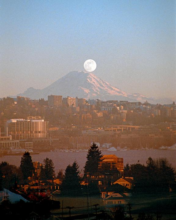 A full moon rises over a snow-capped mountain behind a city and body of water.