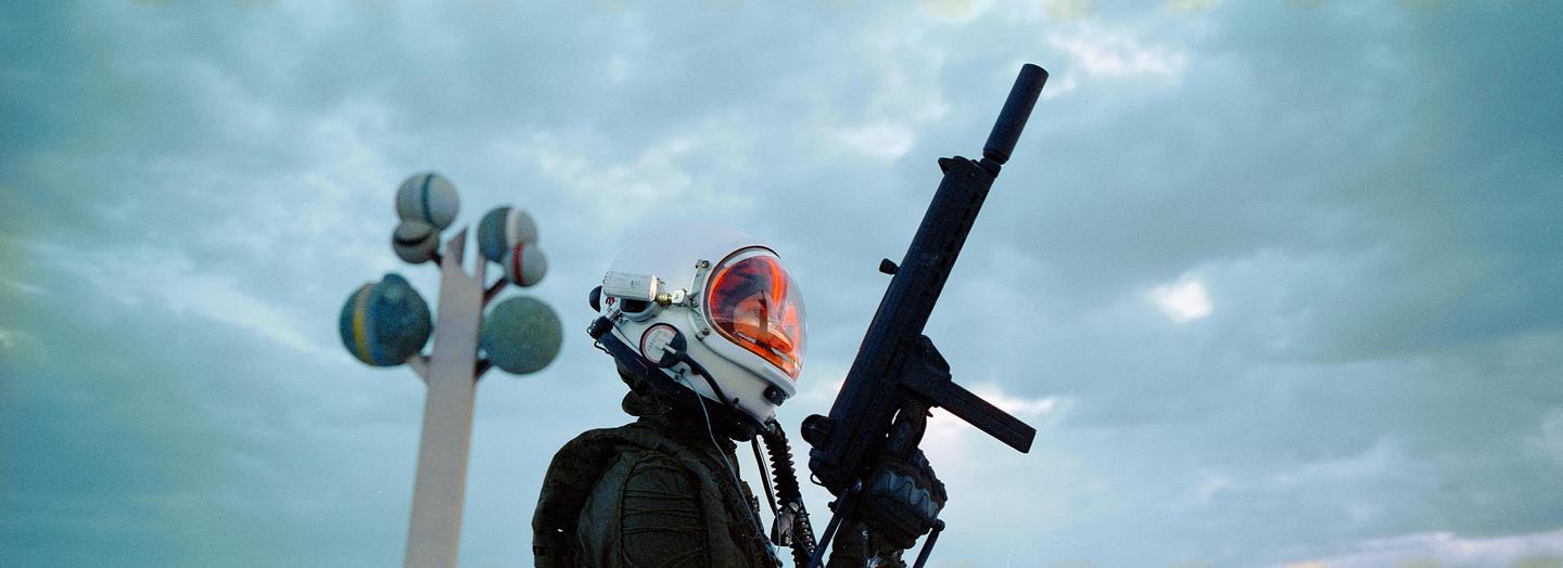 A person in a space helmet holds a rifle with a silencer against a cloudy sky, with a sculpture of spheres on a pole in the background.
