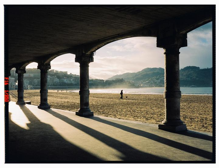 A person walks a dog on a sandy beach with mountains and a city in the background, viewed from under an arched promenade.