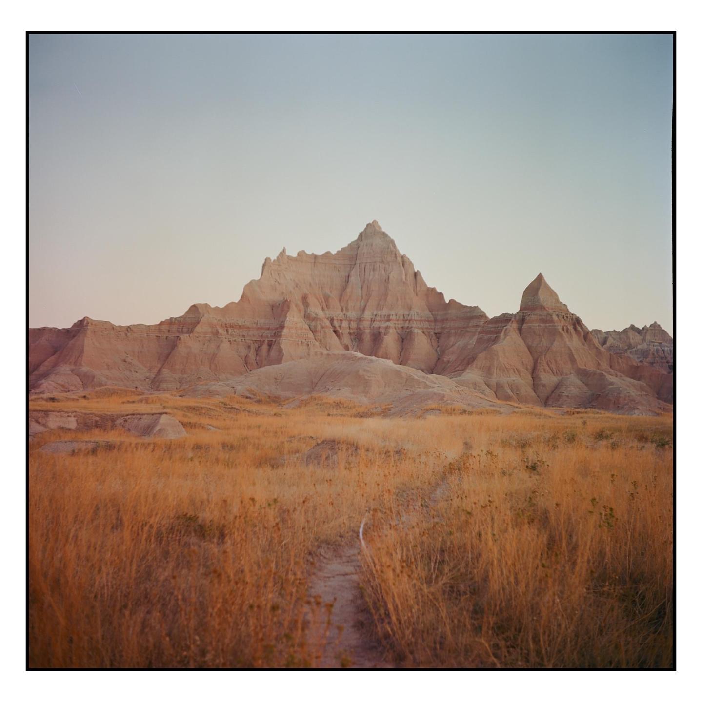 A dirt path leads through a field of dry grass towards a range of rugged, light-colored mountains under a pale sky.