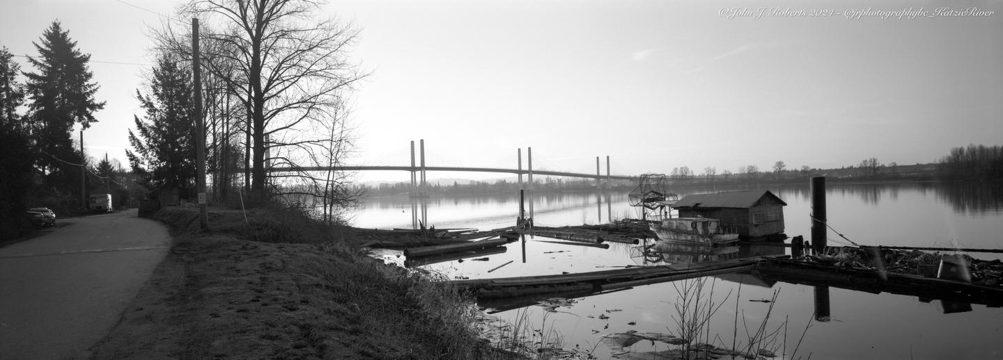 A panoramic view shows a river with a bridge in the distance, a small wooden shack on the water, and a road with trees on the left.