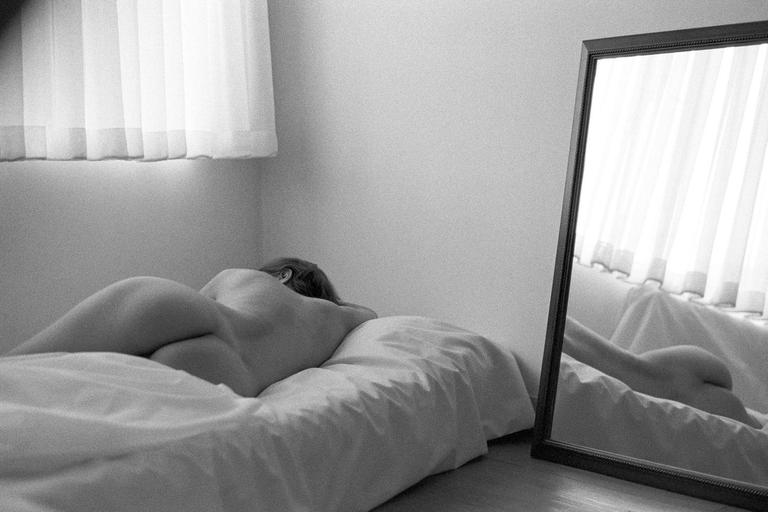 A woman lies on a bed with her back to the viewer, with her reflection visible in a mirror.