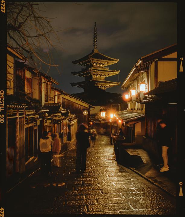 A pagoda rises above a narrow, wet street lined with traditional buildings and people at night.