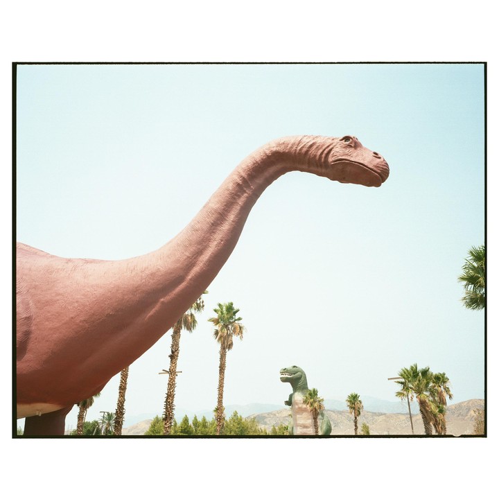 A large dinosaur statue stands in the foreground with palm trees and mountains in the background.