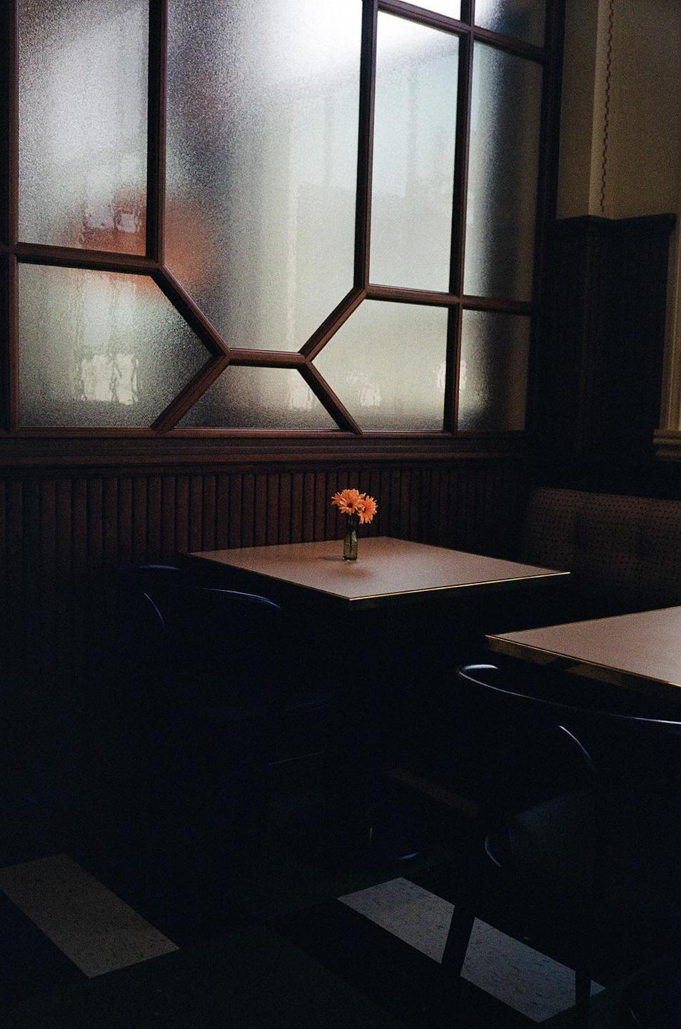 A small vase with orange flowers sits on a table in a dimly lit room with a large window.