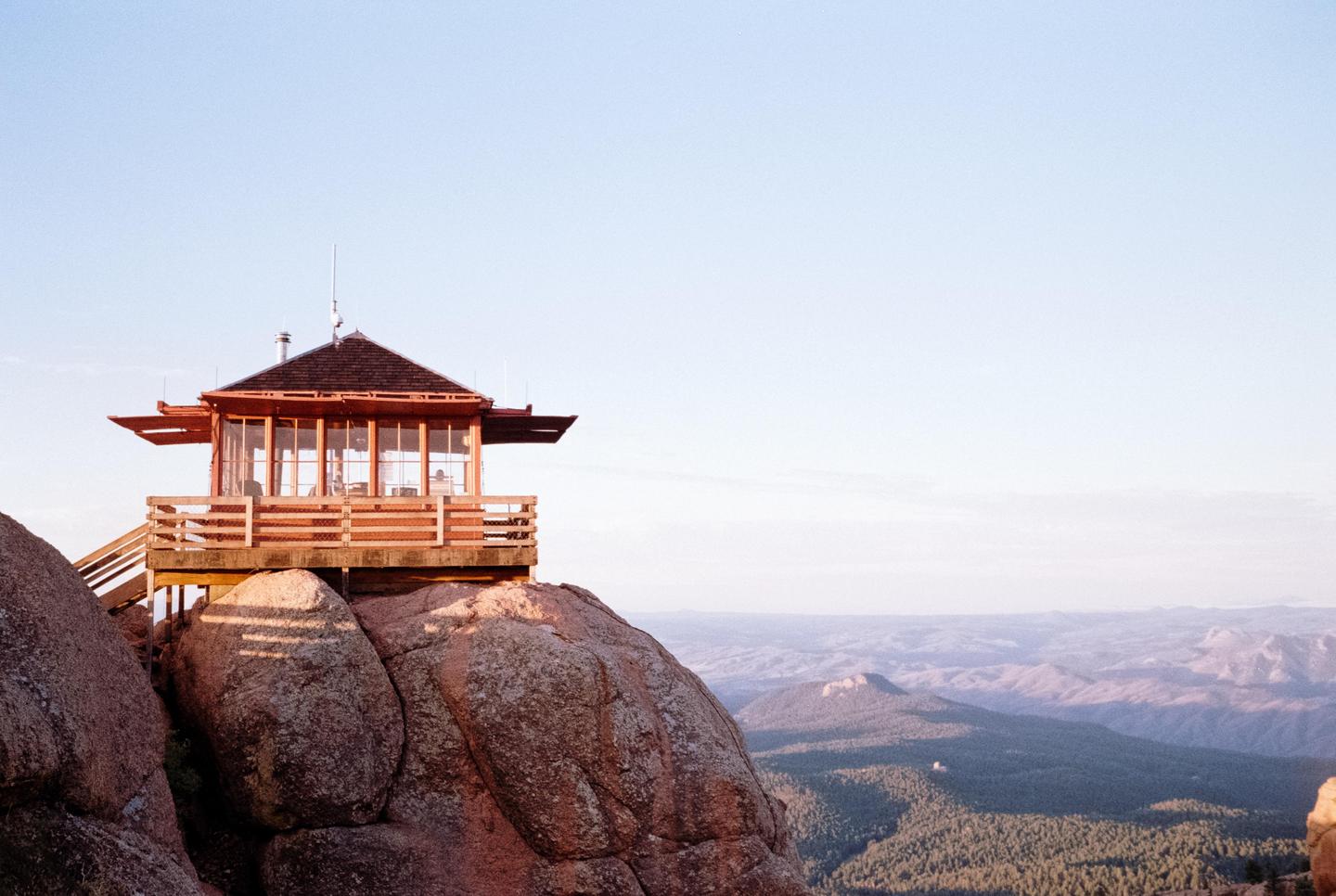 A fire lookout tower sits atop a rocky peak overlooking a vast forest and mountain range.