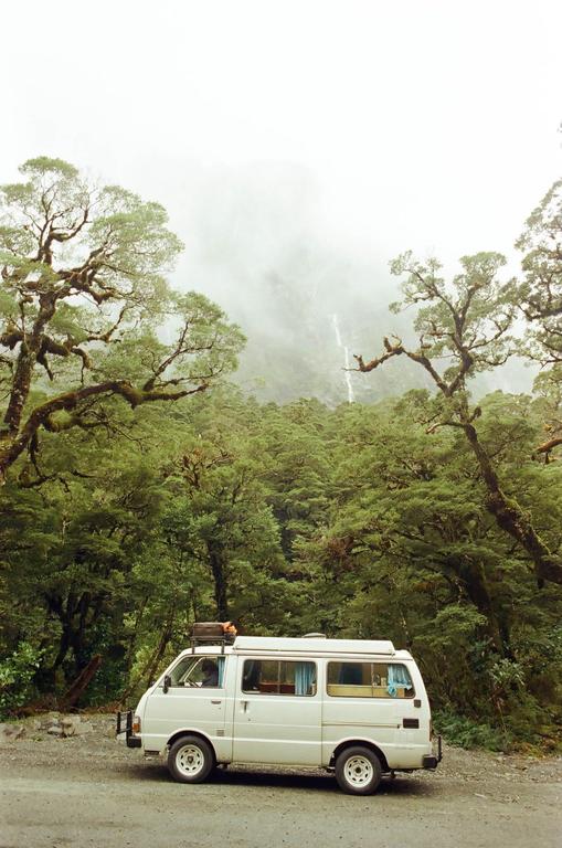 A white van is parked on a dirt road next to a dense forest with a waterfall visible in the misty background.