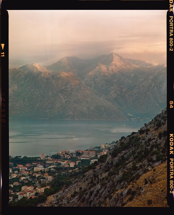 A town with red-roofed buildings sits on a steep hillside overlooking a body of water, with mountains in the background.