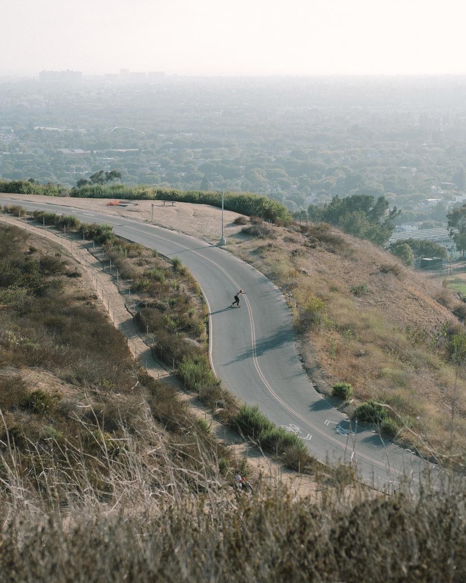 A person on a skateboard descends a winding road on a hill overlooking a hazy city.