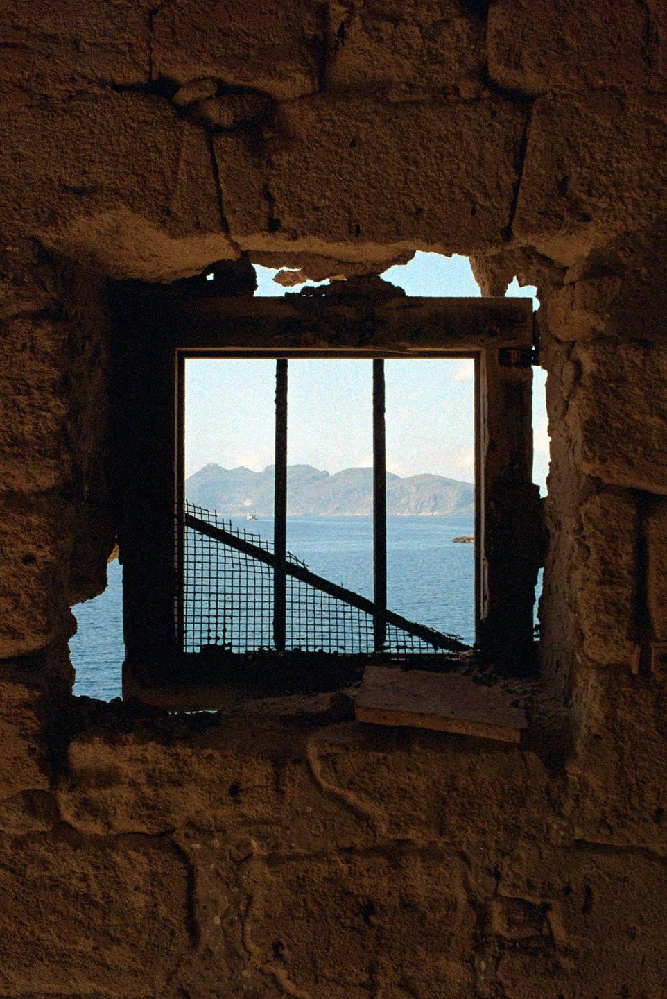 A view of the sea and mountains is visible through a barred window in a stone wall.