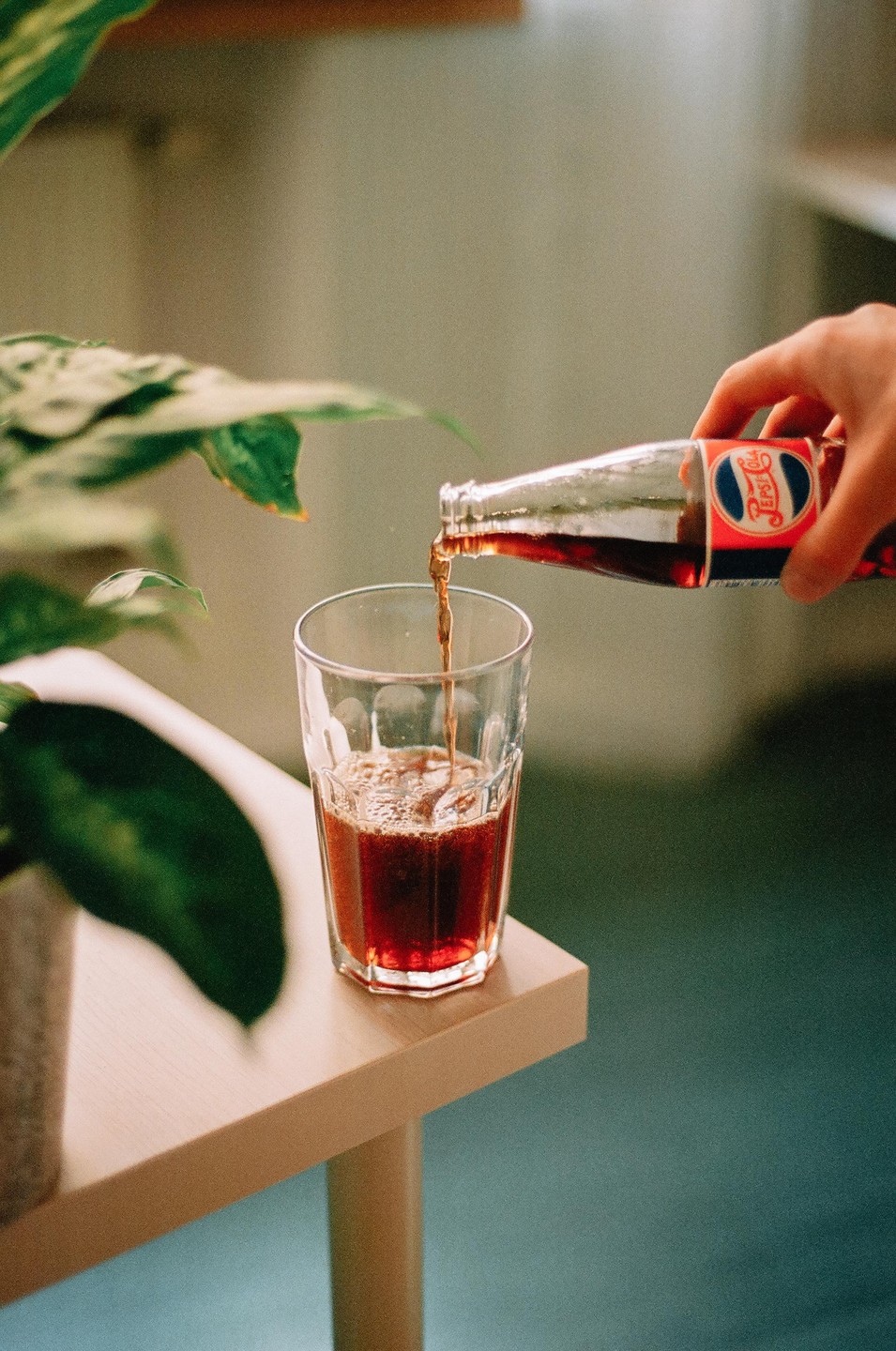 A hand pours Pepsi from a glass bottle into a glass on a wooden table next to a green plant.
