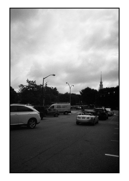 Cars are stopped at an intersection with a church steeple visible in the distance under a cloudy sky.