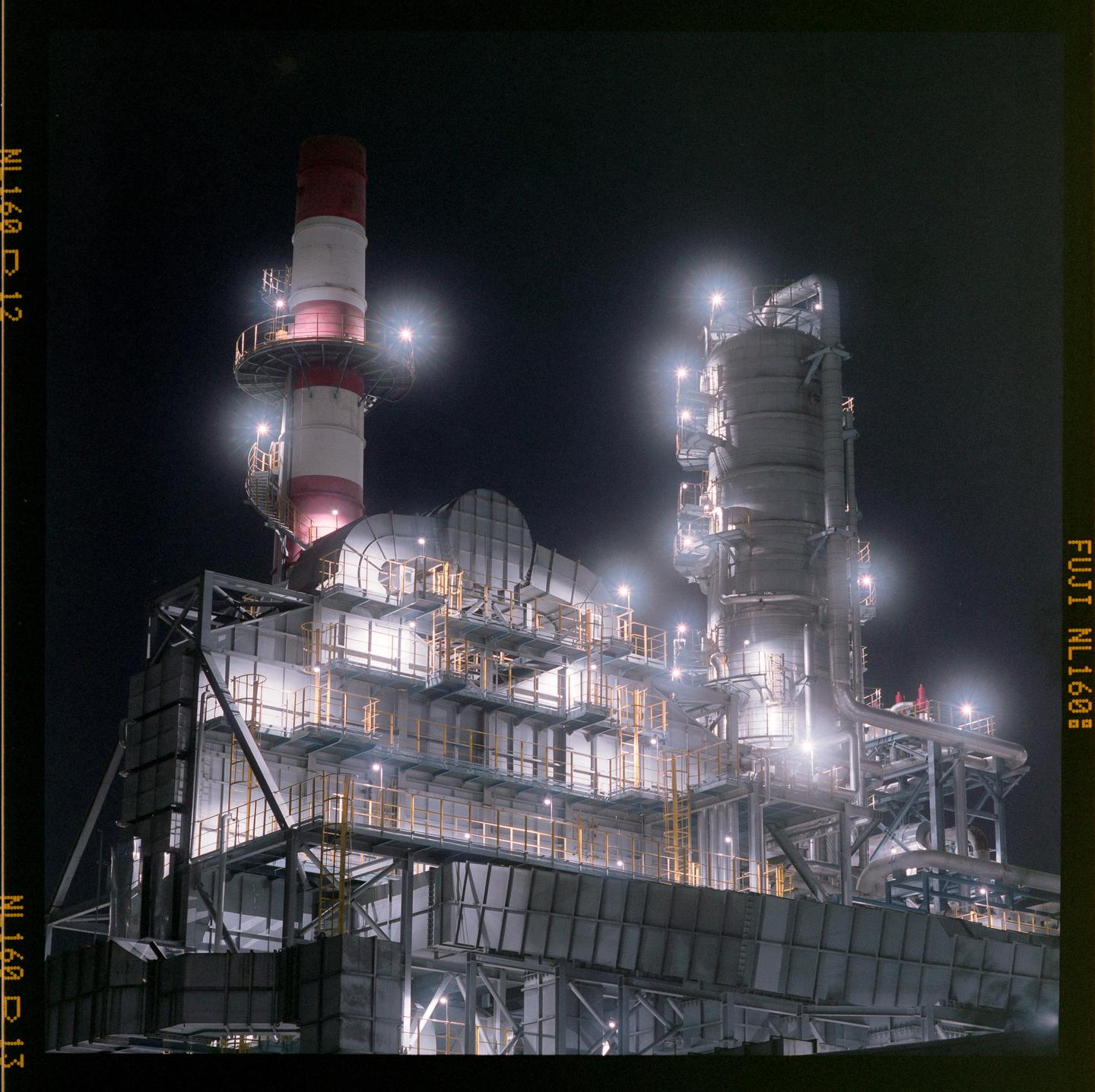 An industrial complex with a tall red and white striped smokestack is brightly lit against a dark night sky.