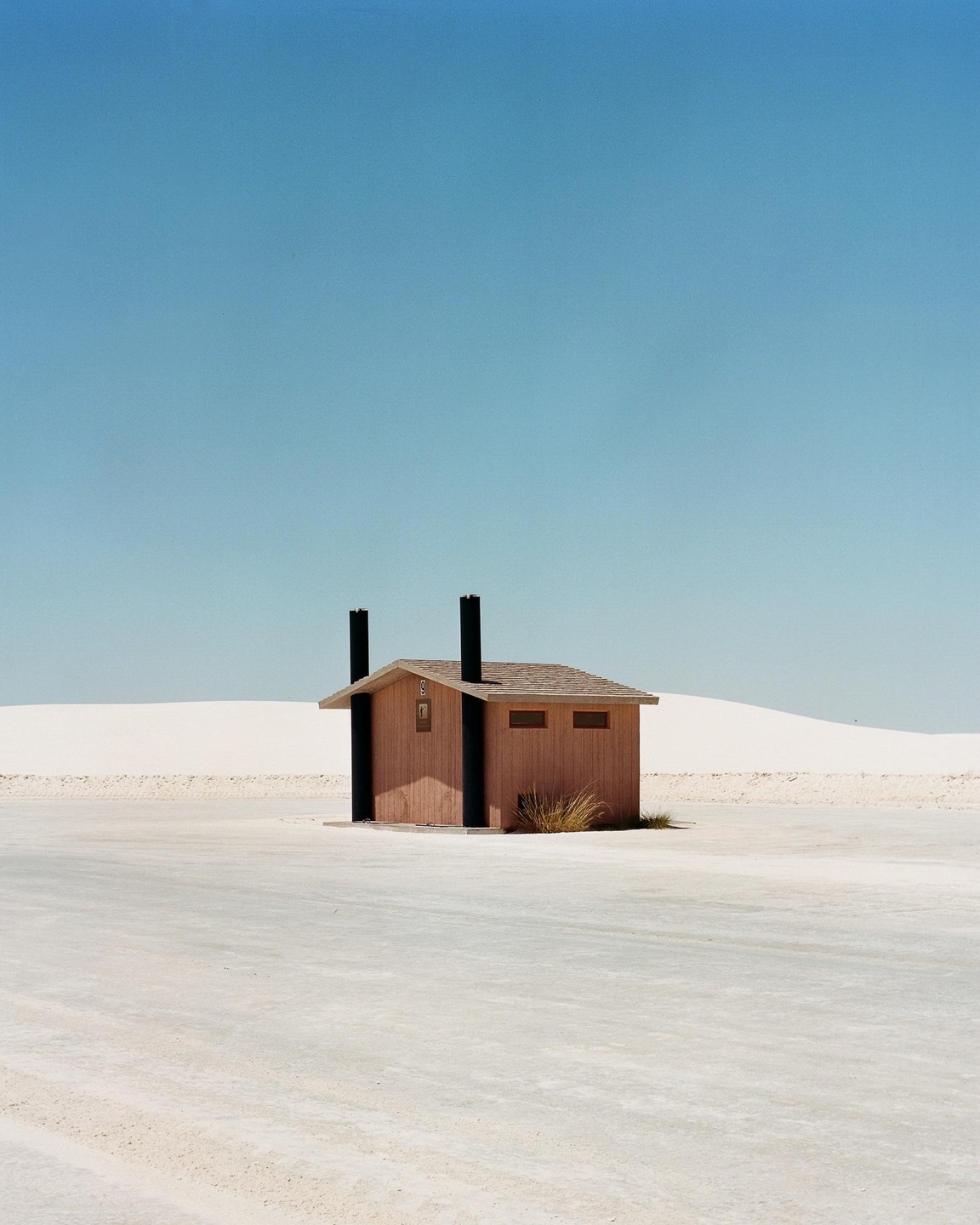 A small wooden building with two black pipes on its roof stands in a vast white desert landscape under a clear blue sky.