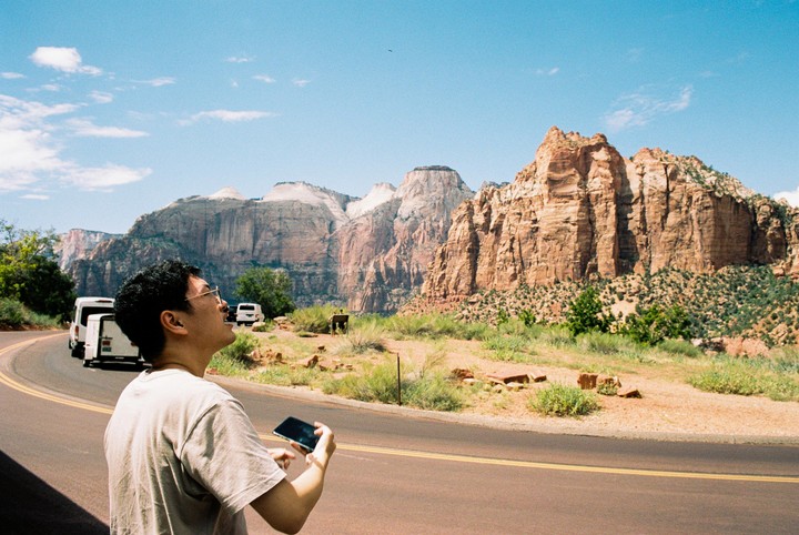 A man stands on the side of a road, looking up at large rock formations under a clear sky.