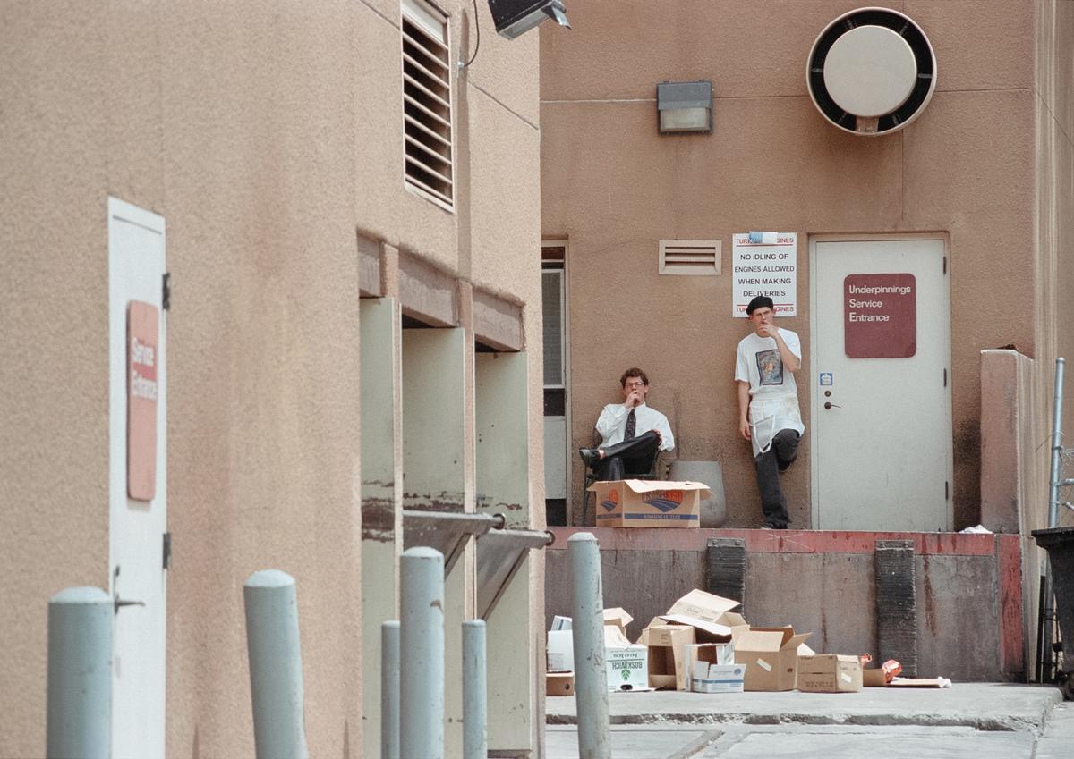 Two men are taking a smoke break outside a service entrance, surrounded by cardboard boxes.