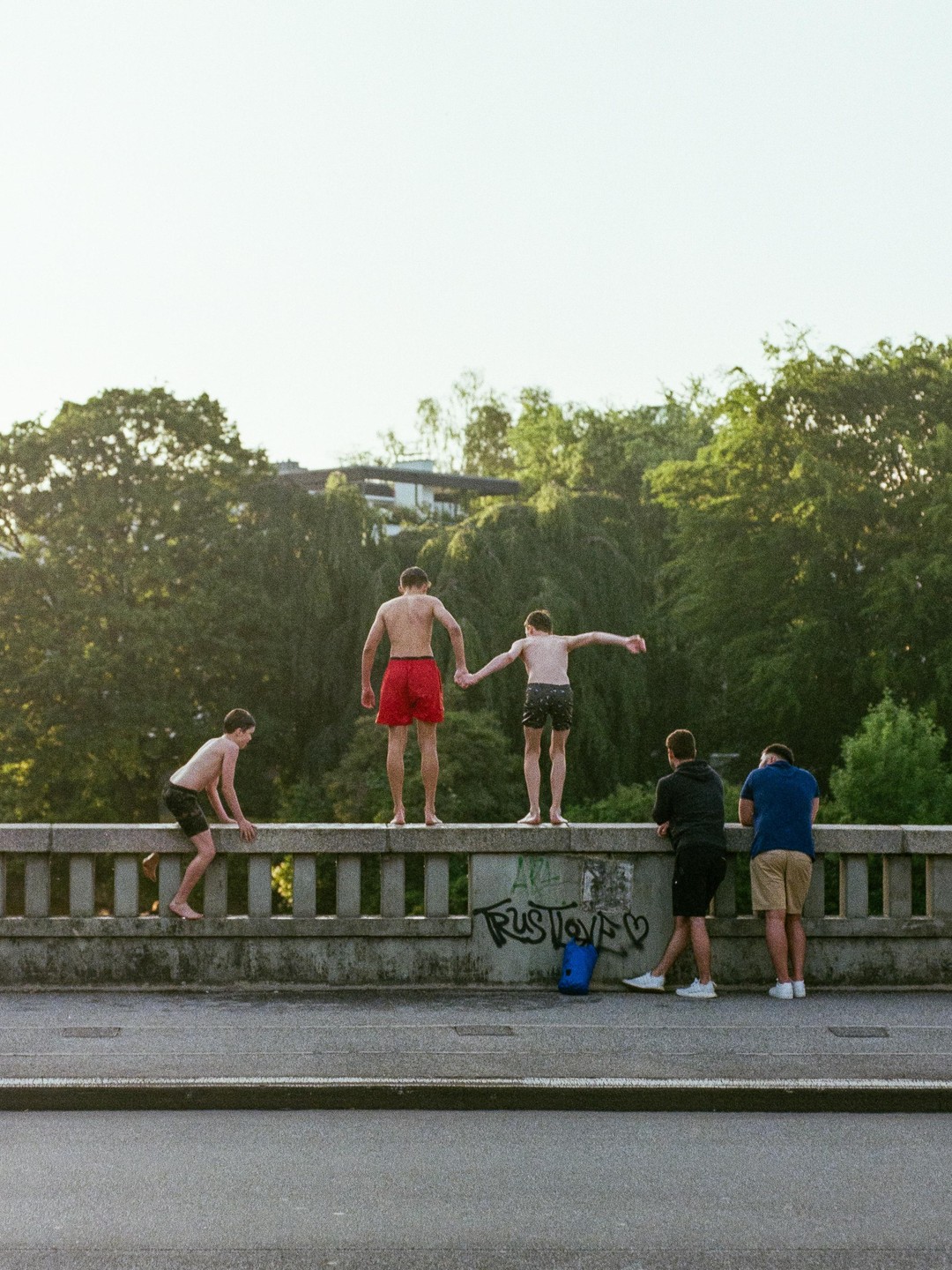 Five boys are on a bridge over a road, with three of them standing on the ledge and two watching.