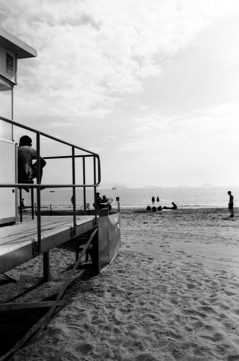 A lifeguard sits on a lifeguard station overlooking a beach with people in the water and on the sand.