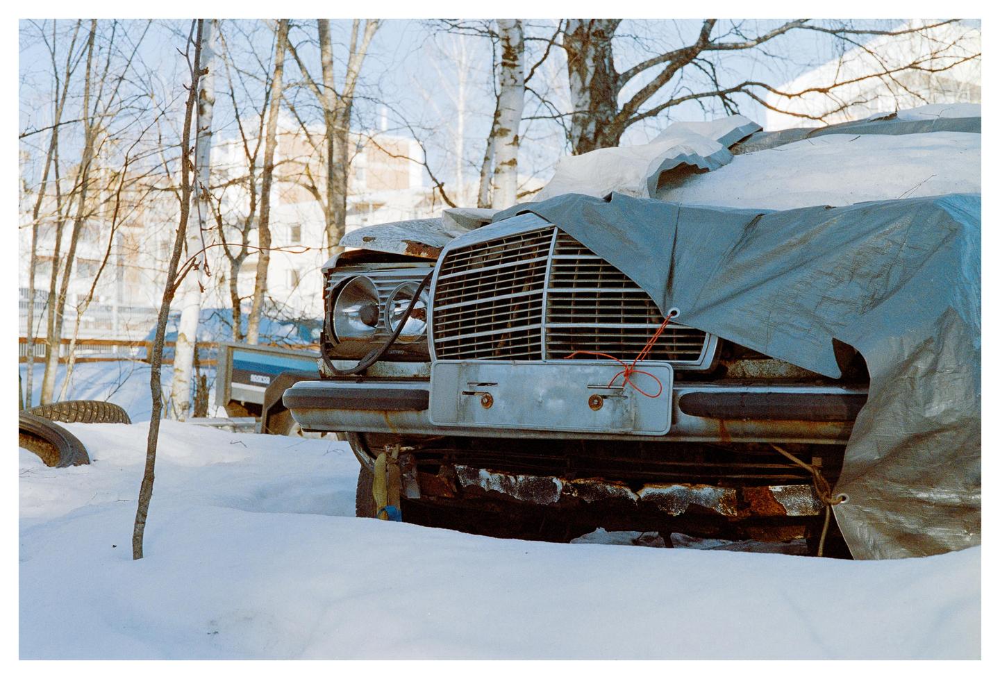 A rusty car covered with a tarp is parked in the snow with trees and buildings in the background.