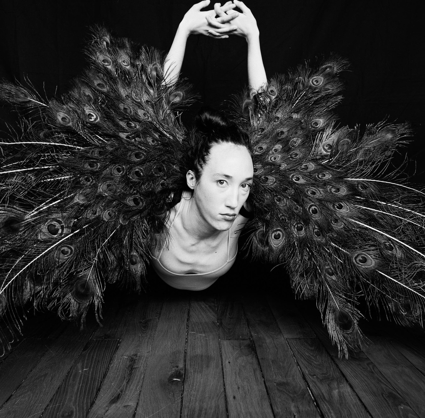 A woman with peacock feathers around her head and shoulders lies on a wooden floor with her arms raised above her head.