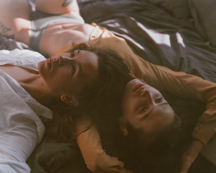 Two women lie on a bed, one in a white shirt and the other in a brown top, with light falling across them.