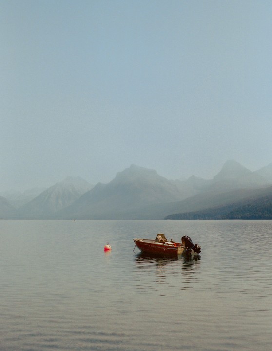 A small red boat floats on a calm lake with hazy mountains in the background.